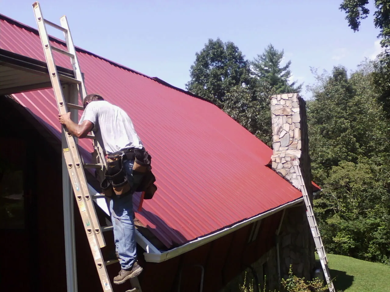 Roofer on a ladder beside a red metal roof and stone chimney — Soffit & Fascia Repair by Roofing Brothers in Woodcliff Lake